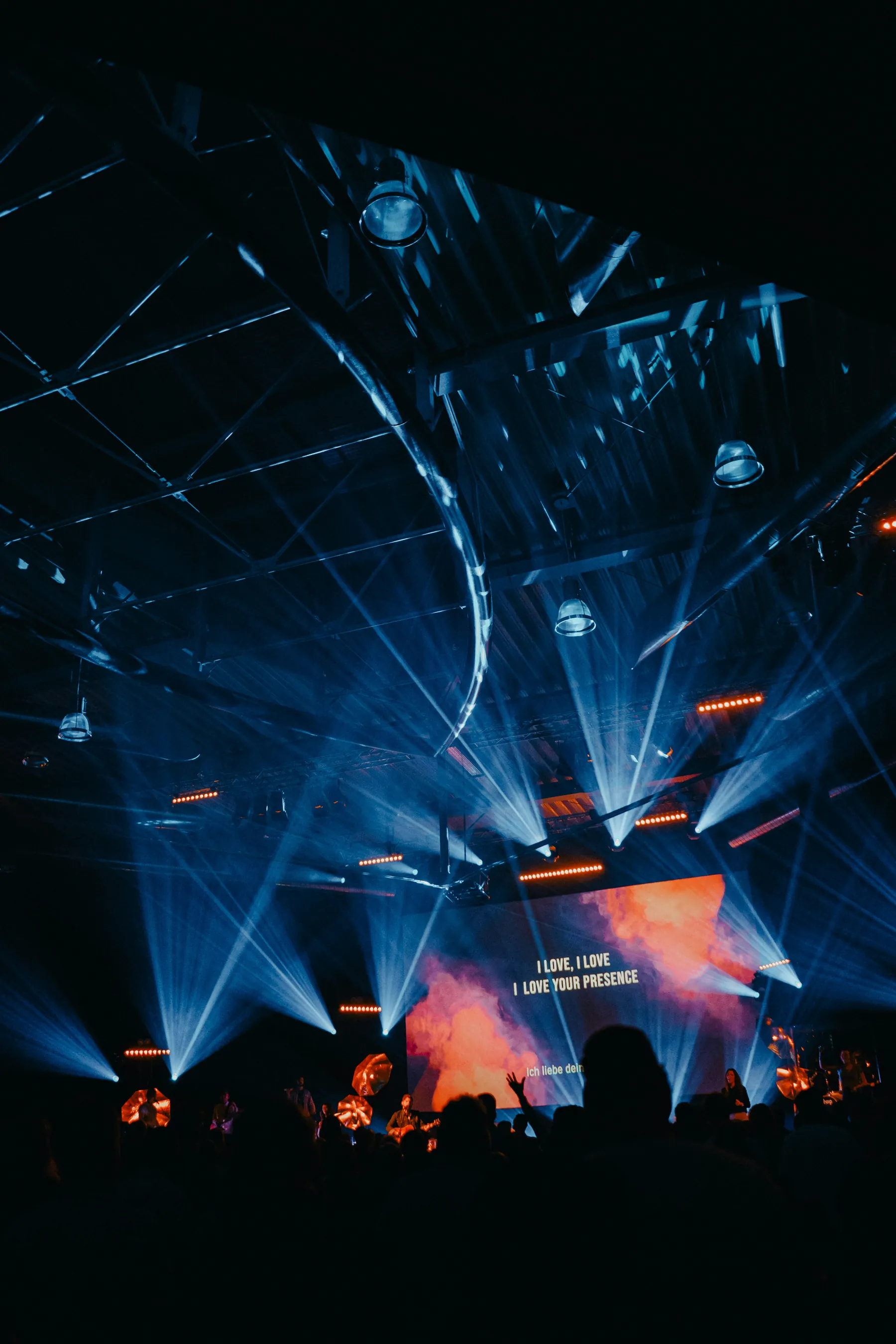 Lighting rigs and beams filling a darkened venue above a large stage screen and a standing audience