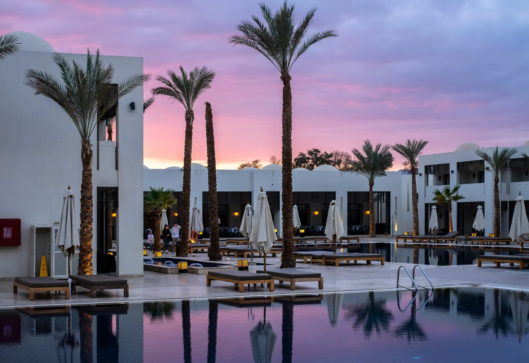 A resort poolside at dusk with palm trees and lounge seating, framed by a pink and violet sunset sky
